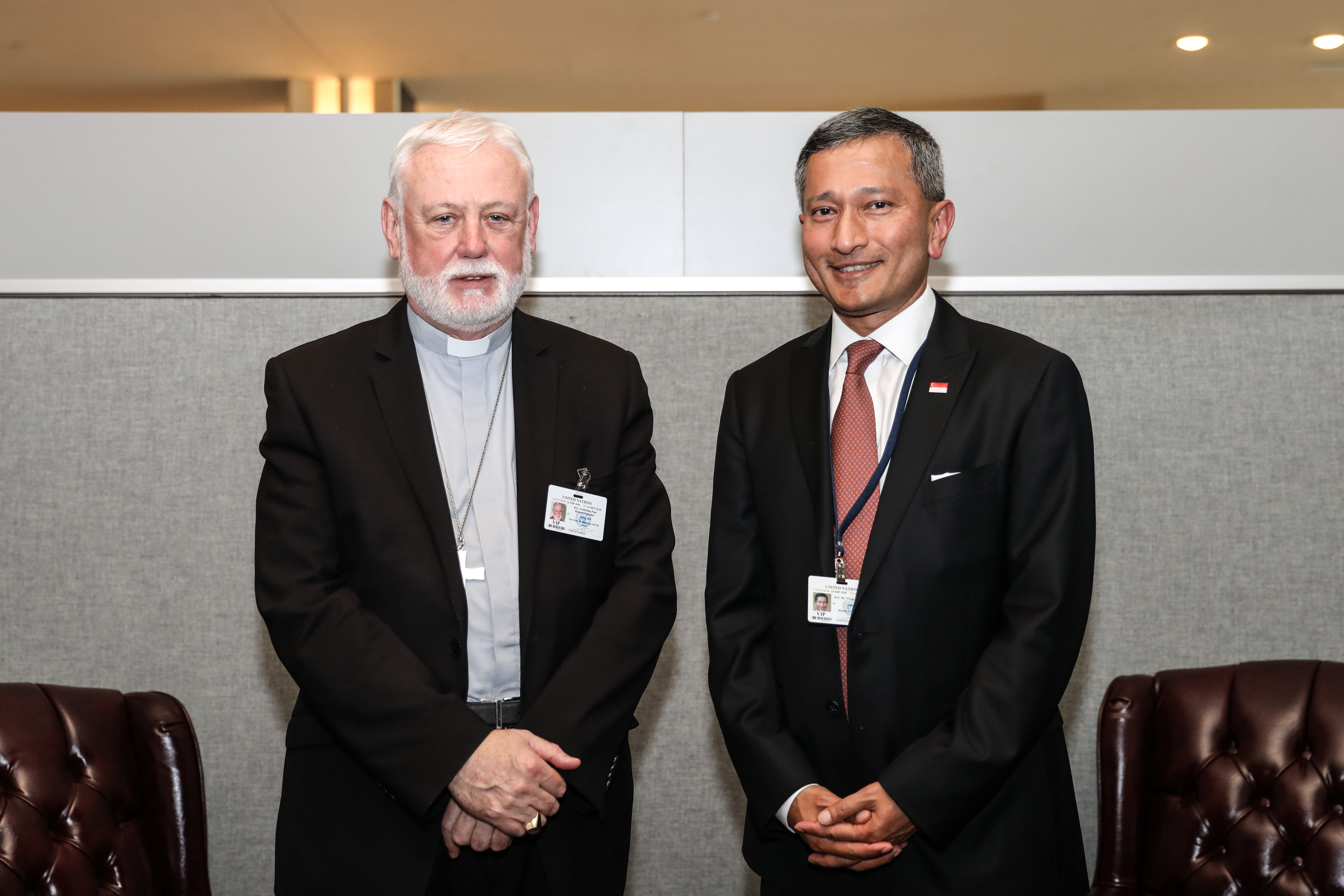 Two men in suits stand side by side. The man on left wears a clerical collar. Both wear ID badges.